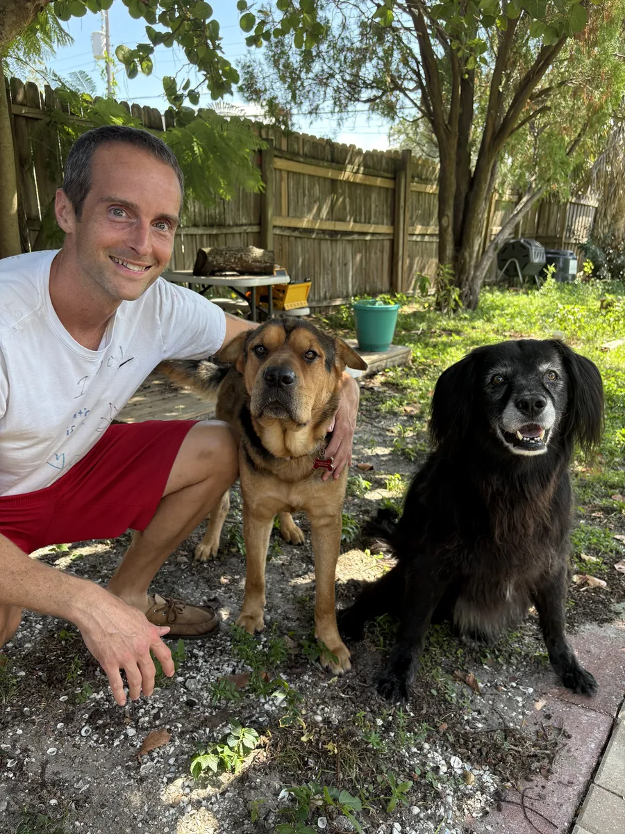 Jerry Banfield crouching in his backyard next to two dogs, one tan and one black