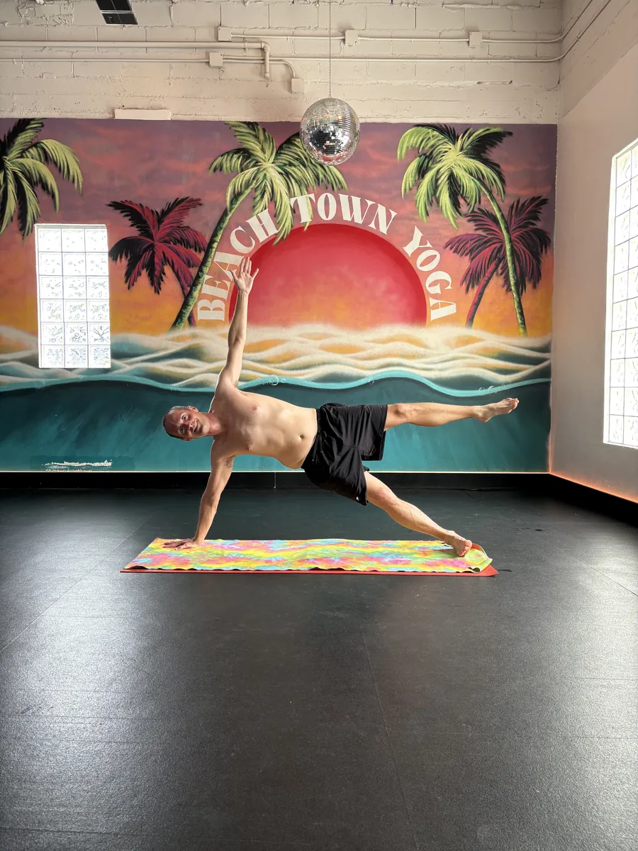Jerry Banfield holding a side plank on a yoga mat in front of a Beach Town Yoga sunset mural