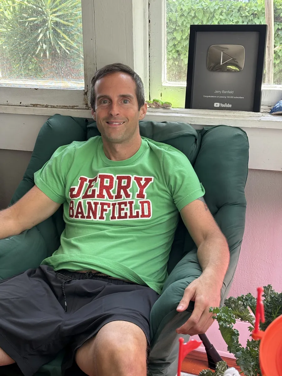 Jerry Banfield in a green armchair wearing the green T-shirt with JERRY BANFIELD in red glitter letters, his YouTube 100,000-subscriber plaque behind him