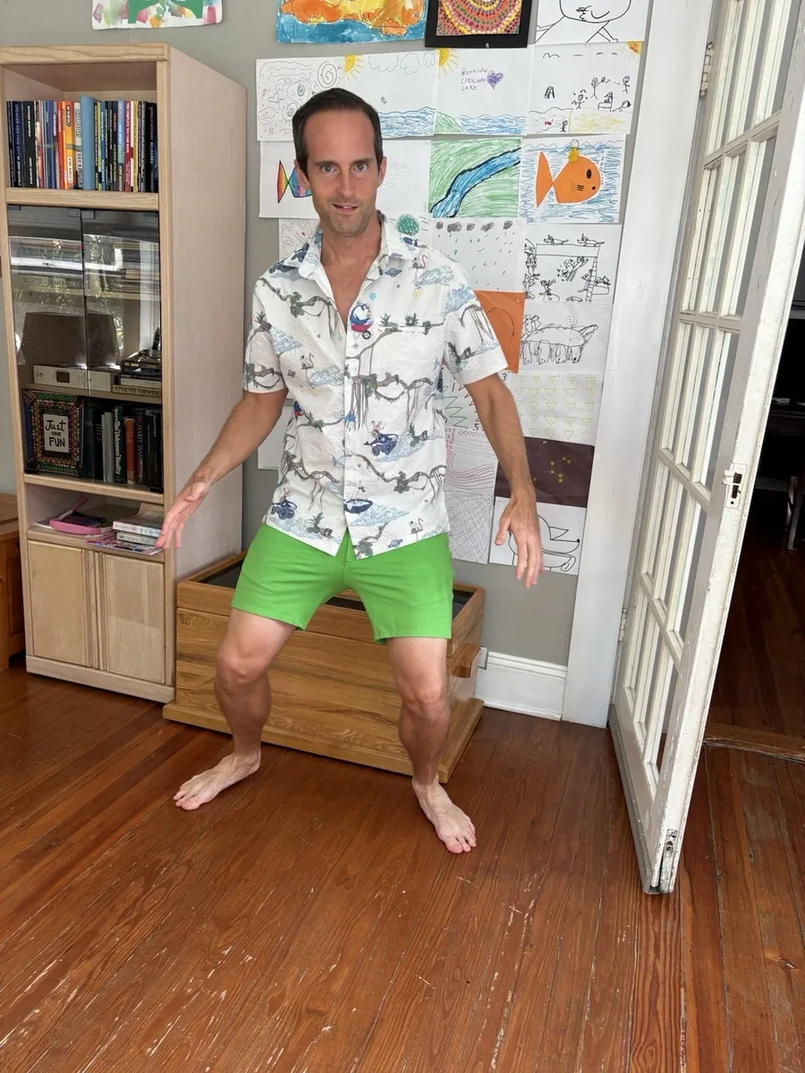 Jerry Banfield standing barefoot on a wood floor in the cream illustrated-print shirt and bright green shorts, children’s drawings on the wall behind him