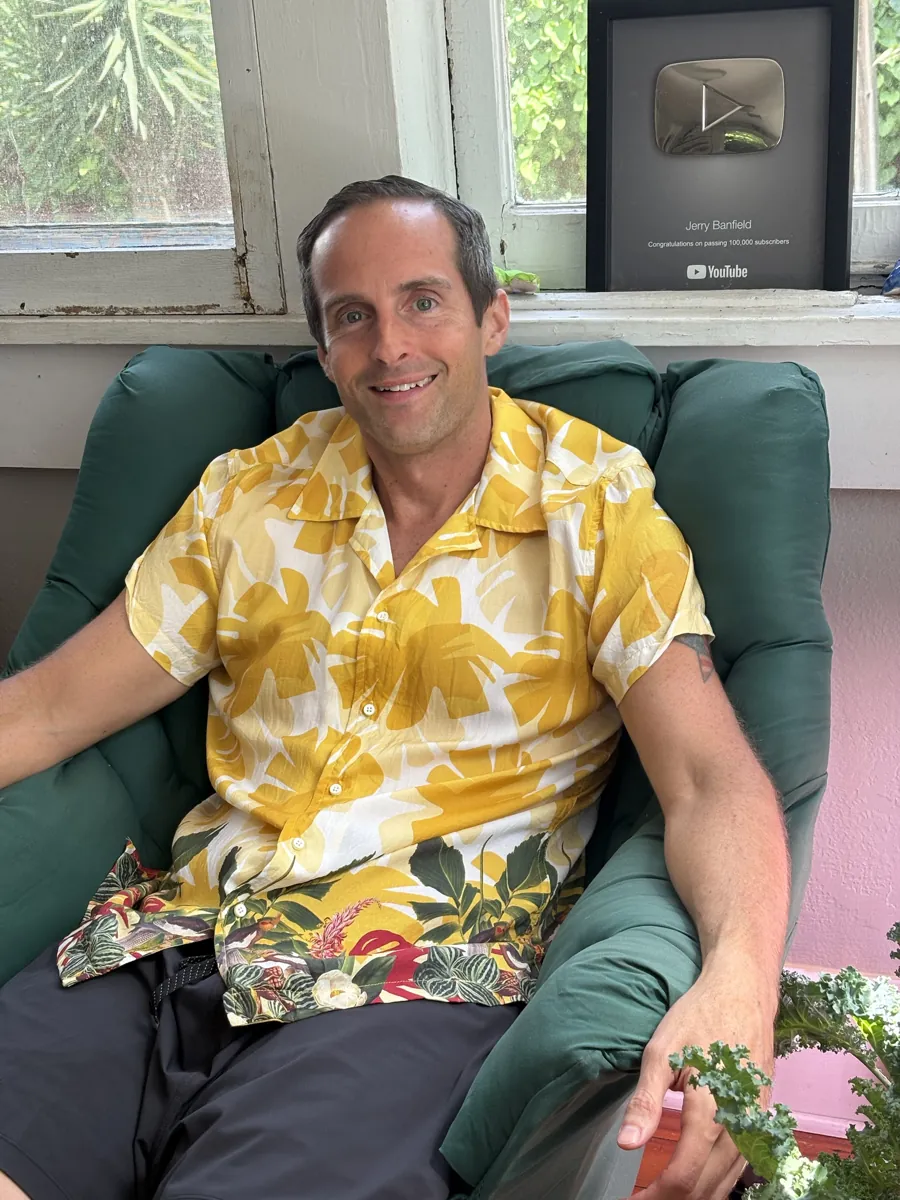 Jerry Banfield smiling in a green armchair wearing the yellow tropical-print shirt, his 100,000-subscriber YouTube plaque on the windowsill behind him